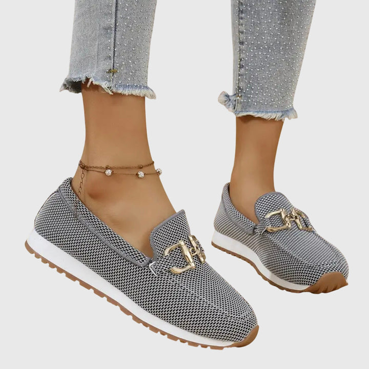 A person wearing white orthopedic loafer sneakers, styled with casual trousers, walking on a paved pathway.